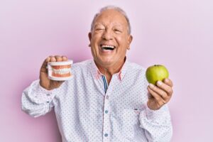 Smiling man holding artificial teeth in one hand and apple in the other